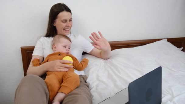 Sorrindo Jovem Mãe Segura Bebê Filha Sentar Cama Acenando Mãos — Vídeo de Stock