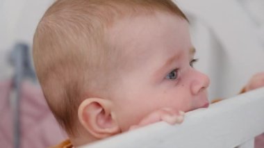 Beyaz beşiğin kenarını çiğneyen tatlı bir kızın seçici odak noktasını kapat çünkü ilk dişi uzayan güzel, oyuncu çocuk rahat yatakta el ele tutuşur. Tıp, pediatri, dişçilik, yeni doğanlar konsepti