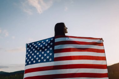 Back view close up of smile young woman waving national USA flag outdoors over sunset sky at summer in mountains, free and happy. American flag, country, patriotism, independence memorial day 4th July