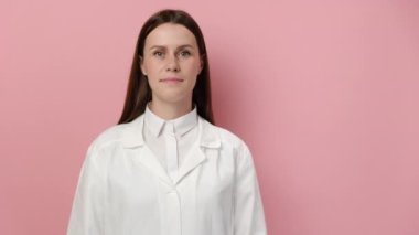 Kendine güvenen, gülümseyen kadın doktor beyaz tıbbi ceket giyer, steteskop pembe stüdyo arka planında duran kameraya bakar. Genç kadın sağlık uzmanı doktor portresi.