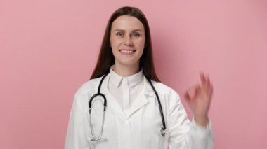 Doktor iyi olduğunu söylüyor. Pembe stüdyo arka planında gülümseyen kadın doktor kameraya bakıyor ve el hareketleriyle tamam işareti gösteriyor, üniforma ve steteskop giyiyor. İnsanlar duygusal yaşam tarzı konsepti