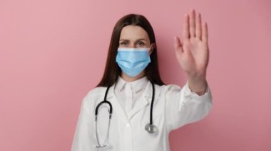 Profesyonel medikal beyaz önlüklü, koruyucu maskeli ciddi profesyonel bir doktor kadının portresi dur işareti ile reddeden davranışları pembe stüdyo arka planında izole edilmiş poz veriyor.