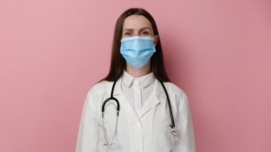 Profesyonel tıbbi maske ve beyaz önlüklü ciddi kadın doktor portresi reddeden baş hareketi, onaylamayan işaret, pembe stüdyo arka planında izole edilmiş poz. Anlaşmazlık kavramı