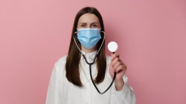 Stetoskop kullanan, profesyonel tıbbi önlüklü ve koruyucu maskeli ciddi bir doktor. Pembe stüdyo arka planında izole edilmiş. Hemşire aletini hasta bir hastayı dinliyormuş gibi kameraya doğrultuyor.