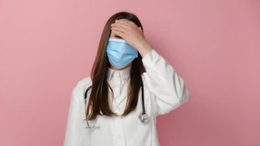 Koruyucu tıbbi maske takmış, yorgun, üzgün kadın doktorlara yakın, alnına dokunan, baş ağrısından bitkin düşmüş terapist doktorlara pembe stüdyo arka planında izole edilmiş pozlar veren, acıyı hisseden,