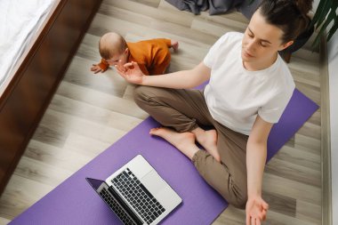 Dikkatli bir anne meditasyon yaparken canlı TV pilates sınıfı özel dersi izlerken dizüstü bilgisayarını kullanarak evde küçük bir bebeğin yanında oturup yoga, sanal eğitim, egzersiz ve meditasyon yapıyor.
