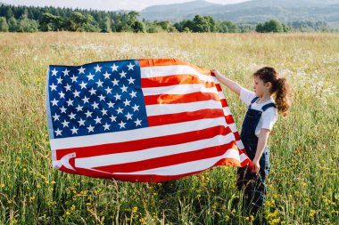 Yeşil çimlerin üzerinde duran sevimli küçük kız çocuğu yaz günü ilham verici ve hayret verici gün batımında ABD bayrağını tutuyor. Bağımsız özgüvenli beyaz çocuk portresi