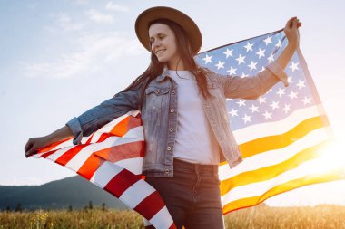 Güzel günbatımında Amerika bayrağı altında arka plandaki yeşil çimlerin üzerinde duran 20 'li yaşlardaki mutlu beyaz kadın. Kendine güvenen, gülümseyen kadın portresi.