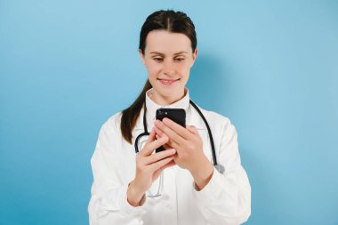 Beyaz önlüklü, gülümseyen tatlı kadın doktor mavi arka planda tek başına duruyor. Akıllı telefonu tut, hastadan gelen mesajları kontrol et, tıp çalışanları için mobil uygulama kullan. Modern teknoloji konseptinin rahat kullanımı.