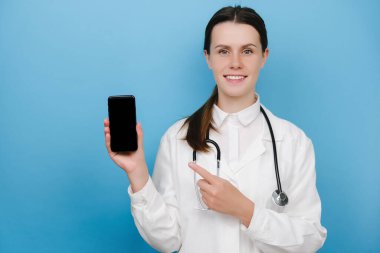 Gezici ekranı işaret eden mutlu genç kadın doktor, kontrolü indirmeyi tavsiye ediyor, danışmanlık başvurusu, mavi arka planda izole edilmiş. Covid-19, sağlık çalışanları ve çevrimiçi tıp konsepti