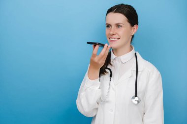 Beyaz tıbbi üniforma giyen gülümseyen kadın doktor ve steteskop elinde telefonla konuşan akıllı telefondan sanal dijital ses asistanı aktifleştir, hastaya internetten danış, mavi arka plan duvarında poz ver.