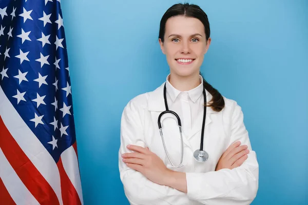 Portrait of happy young woman doctor in white uniform and stethoscope standing near national USA flag , cross arms chest, posing on blue background. Concept of national healthcare system United States