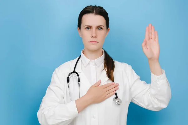 Young caucasian doctor woman wearing medical white coat and stethoscope swearing with hand on chest and open palm, making a loyalty promise oath, posing isolated over blue color background in studio