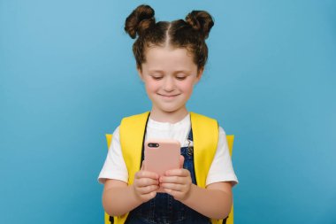Mavi duvarda izole edilmiş mutlu liseli kız. Akıllı telefon. Gülümse, internet uygulamalarını kullan, oyun oyna, çizgi film izle, sarı sırt çantası, modern teknoloji kullanımı kavramı.