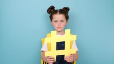 Sırt çantasında sarı hashtag sembolü olan sevimli, sevimli bir kız öğrenci ve kameraya eğlenceli bir şekilde bakıyor. Haşhaş işareti gösteriyor, mavi arka planda izole edilmiş. Moda sosyal medya gönderileri ve okul kavramı