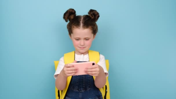 スマートフォンを手にしたバックパックを持った笑顔の女子高生は モバイルアプリを利用したり ビデオゲームをしたり 携帯電話で学んだり モバイル技術のコンセプトで楽しんで 青い背景にポーズをとっています — ストック動画