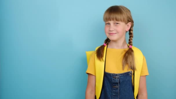 黄色の矢印で脇に示す笑顔の女子高生の肖像は プロモーションコンテンツのためのコピースペースとスタジオで青の色の背景に隔離されたポーズバックパックを身に着けています 学校のコンセプトに戻る — ストック動画