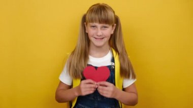 Sevimli sarışın kız elinde küçük kırmızı bir kalp taşıyor sırt çantası takıyor, pastel sarısı arka plan duvarına izole edilmiş ve tanıtım için fotokopi alanı var. Sevgi ve okul konseptine dönüş