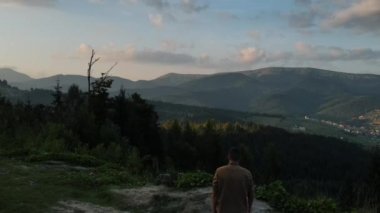 Bin yıllık genç bir adamın günbatımında arka planda yükselen güzel dağ manzarasının insansız hava aracı çekimi. Seyahat konsepti. Çağdaş gezgin doğa yürüyüşü yolunda yürüyor.