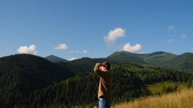 Gezgin genç adam sabah güneşinin altında yeşil güzel dağlara bakıyor. Kahverengi gömlekli sakallı adam çimlerin üzerinde duruyor, sükunetin ve doğanın huzurunun tadını çıkarıyor. Açık hava aktivitesi ve kamp havası.