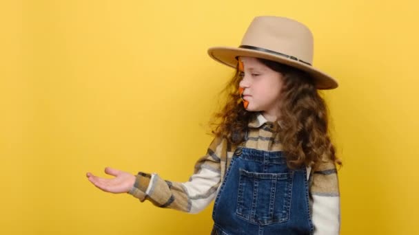 明るい怖いメイクとかわいい女の子の子供の肖像画 プロモーションコンテンツのための黄色のスタジオのコピースペースの脇のポイント カメラを見て笑顔 帽子をかぶって ハロウィンパーティーの場所を示しています — ストック動画