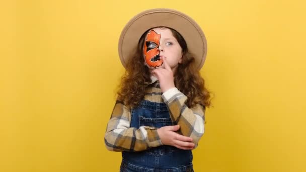 目に見えない小さな女の子の子供の肖像画ハロウィンの化粧マスクは茶色の帽子とシャツを身に着けています周りの考えが出てくる考えを持ち上げ指 黄色の背景スタジオに隔離されたポーズ — ストック動画