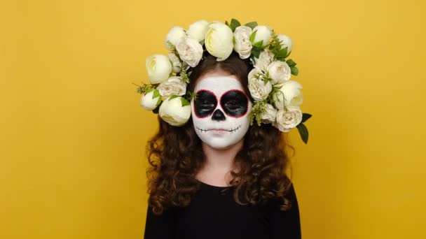 ハロウィンの化粧マスクを持つ明るい女の子の子供の肖像画を閉じます黒の衣装と花の花輪を身に着けています 黄色の背景に隔離された目を閉じて笑顔を保ちます 祝賀会のコンセプト — ストック動画