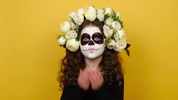 女の子の子供の肖像画は メキシコのカーニバルの準備を頭蓋骨のメイクを着て 黒の衣装や花の花輪に身を包んだ空気のキスを吹くと 黄色の背景に隔離された怖い表情をしています ハロウィンのコンセプト — ストック動画