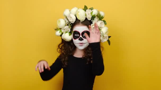 Szoros portré egy kislányról, mexikói cukorkoponyás sminkkel és virágkoszorúval, felemeli a kezét és a kamerába néz, sárga háttérrel elszigetelve. Kreatív, művészi, halloween koncepció