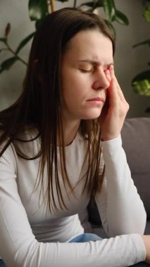 Yorgun, sağlıksız genç bir kadın, evde tek başına oturuyor, gözleri kapalı, elini başını tutuyor, şiddetli baş ağrısı veya migrenden muzdarip, zonklayan ağrı konsepti