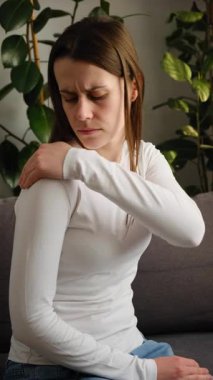 Evdeki kanepede oturan mutsuz, üzgün, omuz ağrısı çeken genç bir kadın. Sağlık ve ilaç. Acı vücut kasları sert problem konsepti