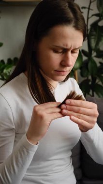 Kırık uçlu, dağınık, kuru saçlı, dağınık saçlı, evdeki kanepede oturan hayal kırıklığına uğramış, endişeli genç bir kadın. Hasarlı saç. Güzellik konseptinin sağlık hizmeti