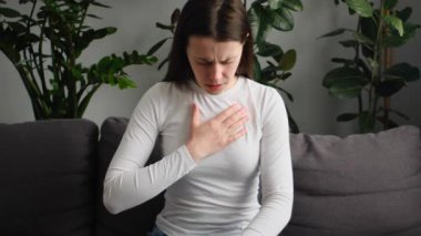 Kanepede tek başına otururken göğüs ağrısı çeken sağlıksız beyaz bir kadın. Üzgün kadın evde kalp ağrısı, damar tıkanıklığı ya da kalp hastalığı konsepti yaşıyor.