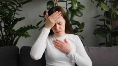 Hasta gripli genç kadın baş ağrısı, alna dokunan el, vücut ısısını ölçmek için termometreyi tutmak, evdeki kanepede dinlenirken hastalanmak. Sağlık hizmeti kavramı