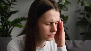 Yorgun, sağlıksız genç bir kadın, evde tek başına oturuyor, gözleri kapalı, elini başını tutuyor, şiddetli baş ağrısı veya migrenden muzdarip, zonklayan ağrı konsepti