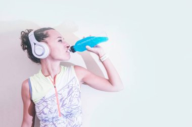 Genç bayan sporcu enerji içeceği içiyor ve kulaklıkla müzik çalıyor.