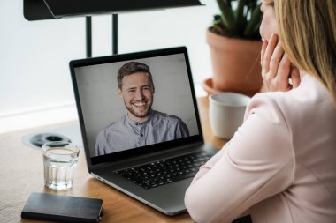 Uzaktan kumandalı online çalışan bir kadın iş masasında laptopuyla oturuyor ve bir online toplantıya katılıyor ya da video konferansı veya webinar sunumu izliyor.
