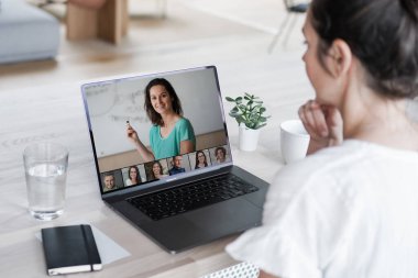 Uzaktan kumandalı online çalışan bir kadın iş masasında laptopuyla oturuyor ve bir online toplantıya katılıyor ya da video konferansı veya webinar sunumu izliyor.