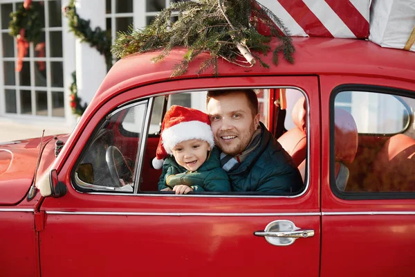 Przystojny mężczyzna z uroczym chłopcem w kapeluszu Świętego Mikołaja wewnątrz czerwonego samochodu retro z prezentów świątecznych — Zdjęcie stockowe