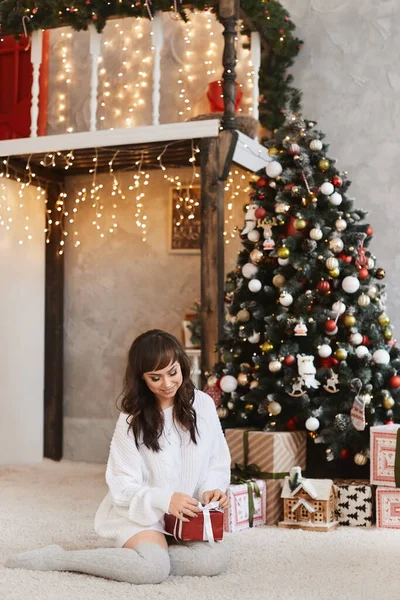 一个年轻美丽的女人，黑发，穿着舒适的衣服，打开圣诞礼物，装饰圣诞的内部。穿着针织连衣裙和长袜的模特在新年的室内装扮 — 图库照片