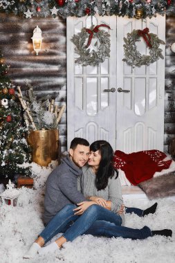 Genç, yakışıklı bir adam ve genç bir kadın, gri kazaklar içinde, kapalı bir Noel ağacının yanında sarılıp poz veriyorlar.