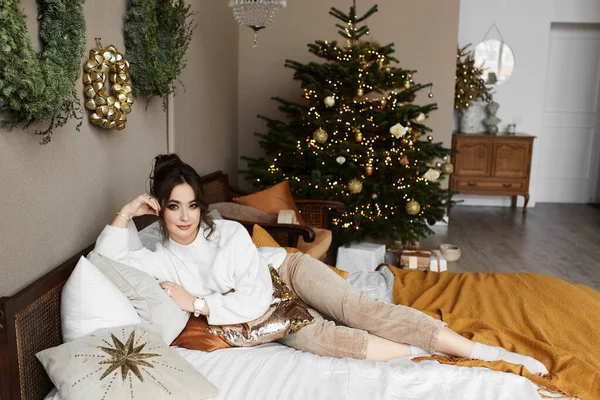 Gyönyörű fiatal nő fényes smink pózol az ágyon, közel a karácsonyfa a hálószobában belső — Stock Fotó
