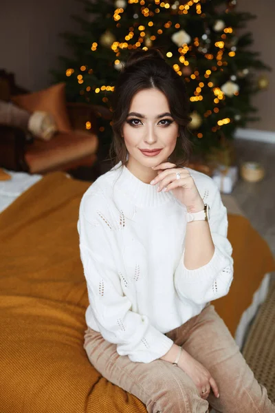 Mooie volwassen vrouw met heldere make-up zit op het bed met feestelijke lichten van een kerstboom op de achtergrond — Stockfoto