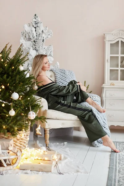 Preciosa mujer adulta en ropa de casa acogedora posando en el sillón vintage cerca del árbol de Navidad decorado —  Fotos de Stock