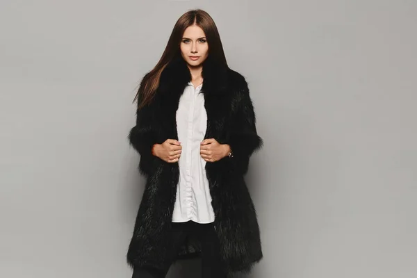 젊은 모델 여성이 회색 배경에 긴 겨울 모피 코트를 입고 밝은 남성성을 띄고 있습니다. 스튜디오 촬영, 겨울 패션 — 스톡 사진