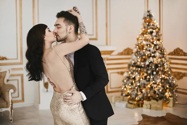 Homem bonito em um terno preto e um jovem modelo em um vestido de noite elegante com costas nuas posando no interior de luxo decorado para o tempo de Natal — Fotografia de Stock