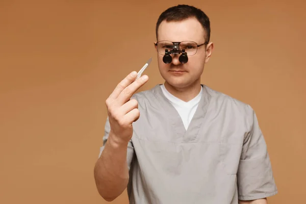 Adult doctor or surgeon man in magnification glasses posing with a scalpel at the beige background, isolated. Focus on the scalpel
