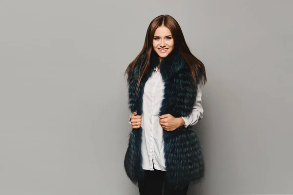 Smiling model girl in winter fur vest posing on the grey background, isolated. Winter fashion