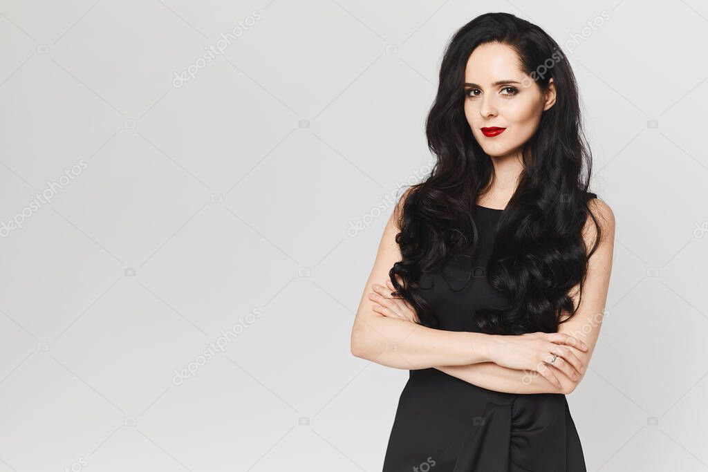 Confident adult woman with bright red lips in a black dress posing with crossed arms and looking in the camera at the white background, isolated with copy space for your logo or advertisement.