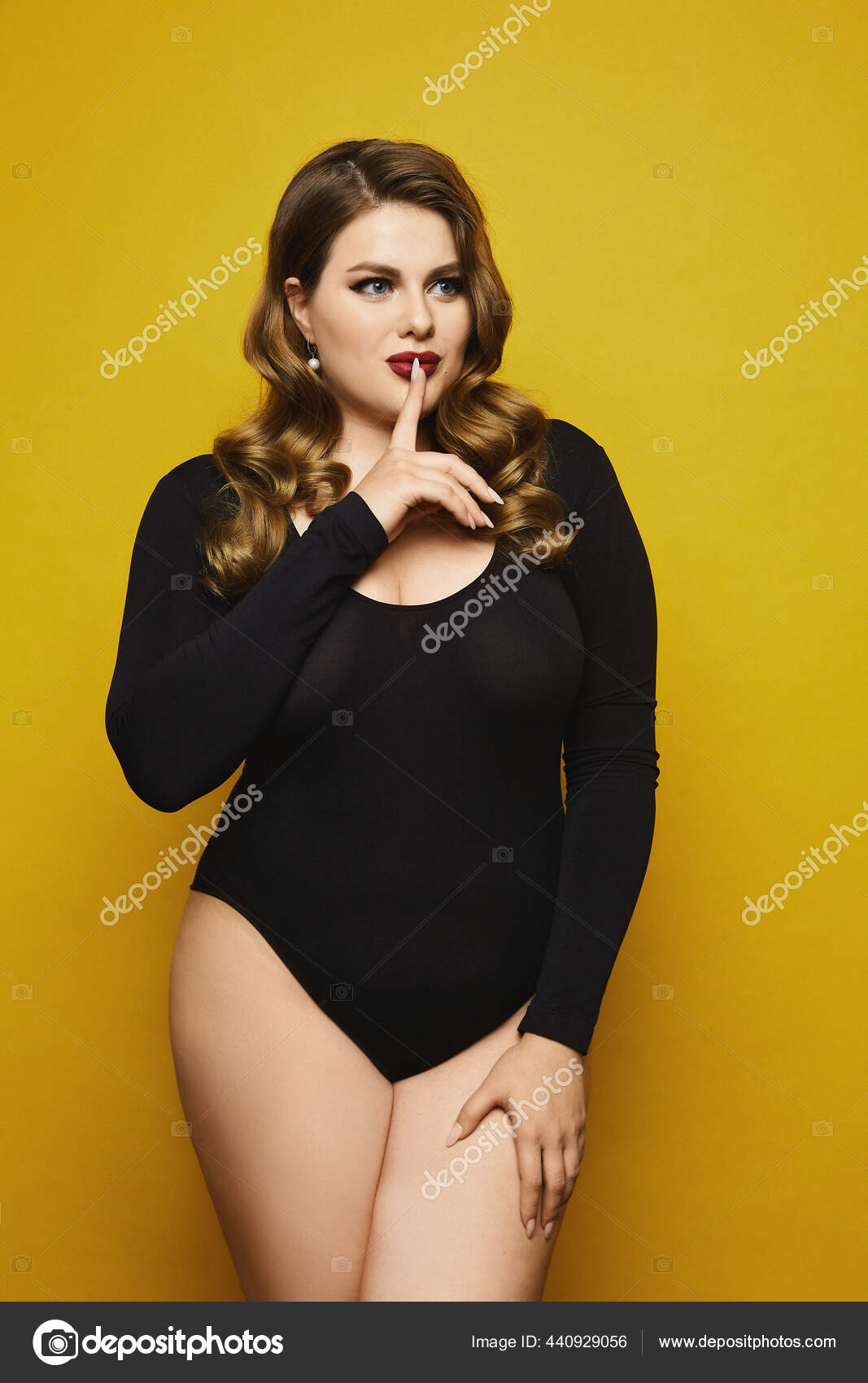 Plus size sexy model girl, with bright makeup, in black bodysuit saying  hush and posing at the yellow background. Young plus size woman in black  bodysuits isolated on the yellow background. Stock